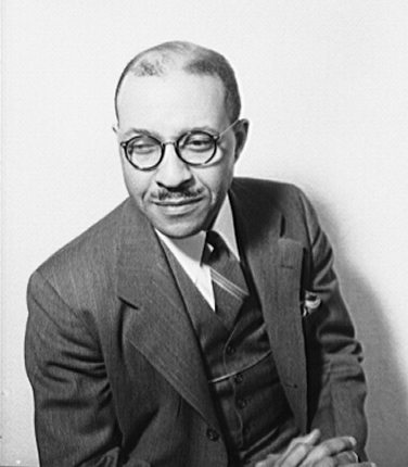 Charles Johnson sits, hands folded, wearing round spectacles.