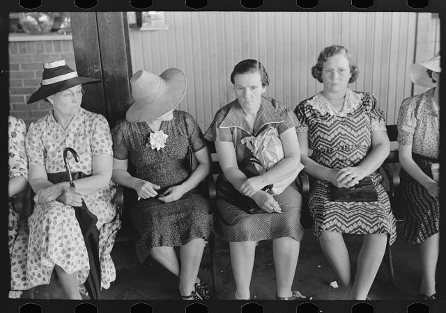 Six women in dresses sit on a bench waiting.