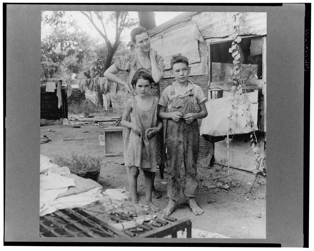 A mother and her two children wearing filthy clothing stand in front of their make-shift home. Clothes hang on the line to dry in he background.