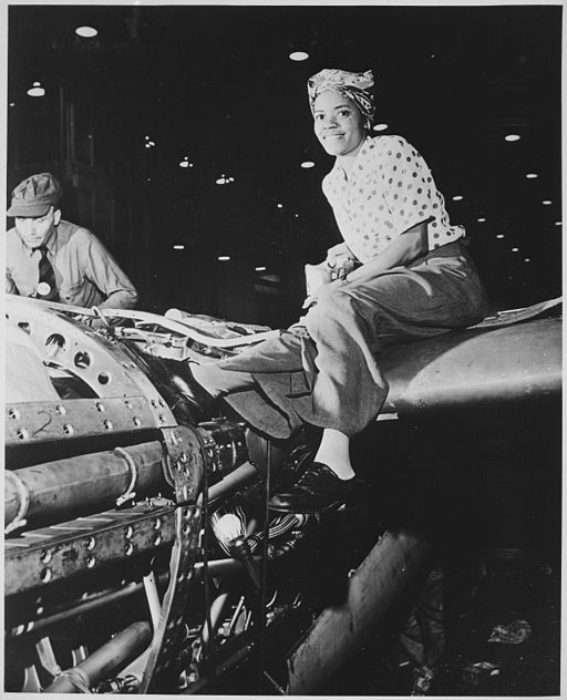 A female, African American worker wearing a head scarf sits on top of the aircraft she is building